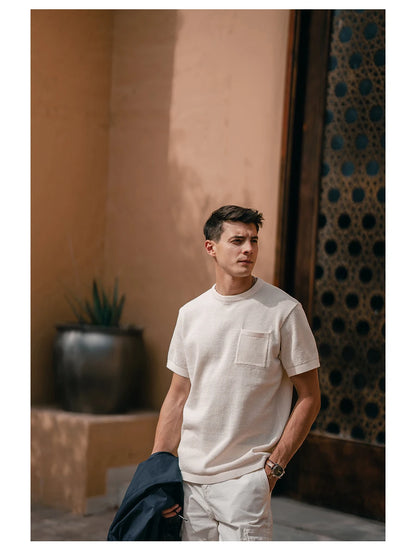 Man wearing a white t-shirt and pants standing against a textured wall with a plant in the background.