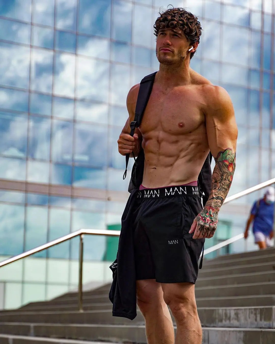 Man with a tattooed arm wearing black shorts outdoors with a modern building in the background
