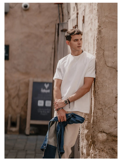 Man leaning against a textured wall wearing a white t-shirt and beige pants.