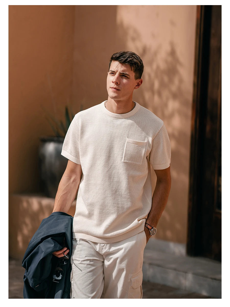 Man wearing a beige t-shirt and pants standing against a warm-toned wall.