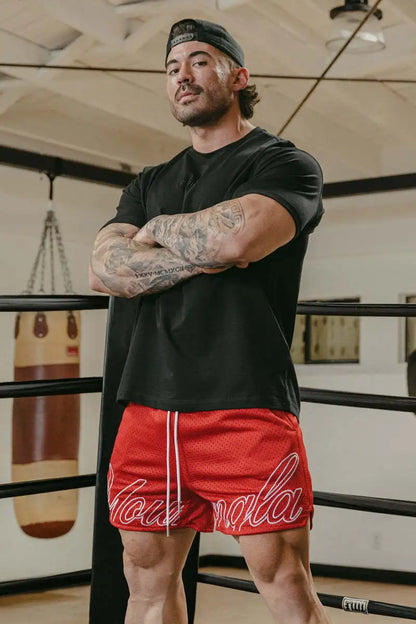 Man in black t-shirt and red shorts with white text standing in a boxing ring.