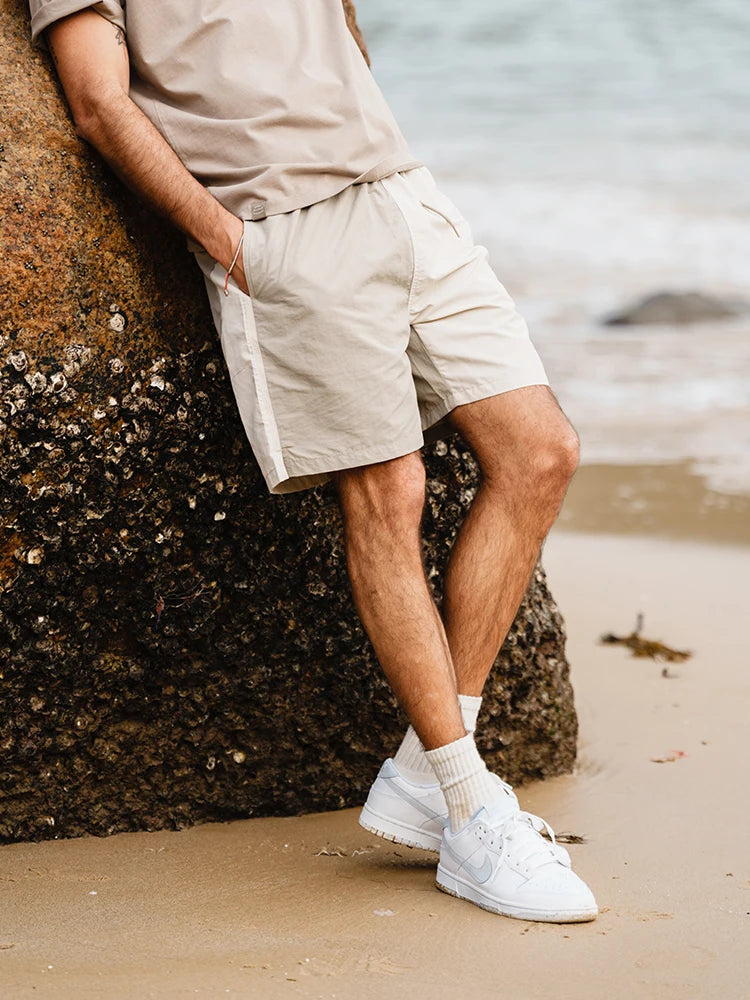 Person wearing beige shorts and white sneakers leaning against a rock on a beach.