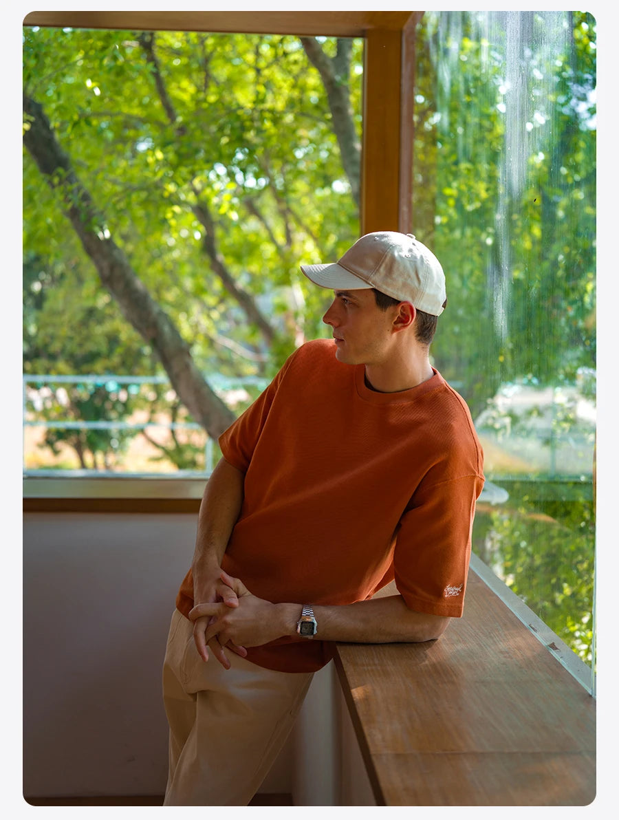 Person wearing an orange shirt and white cap sitting by a window with greenery outside