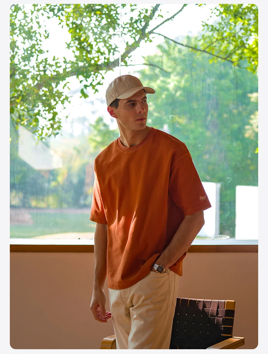 Man wearing an orange shirt and beige cap standing indoors with greenery outside