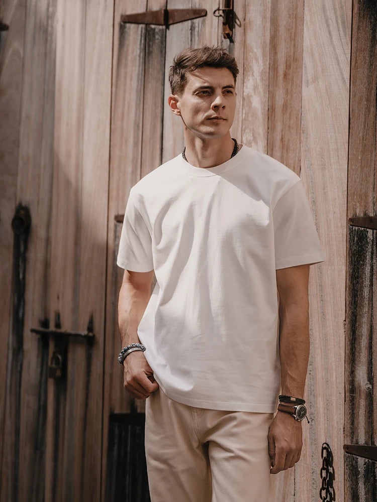 Man wearing a white t-shirt and beige pants standing against a wooden wall.