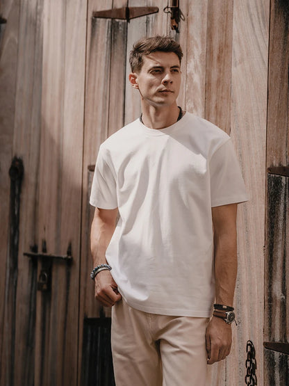Man wearing a white t-shirt and beige pants standing against a wooden wall.